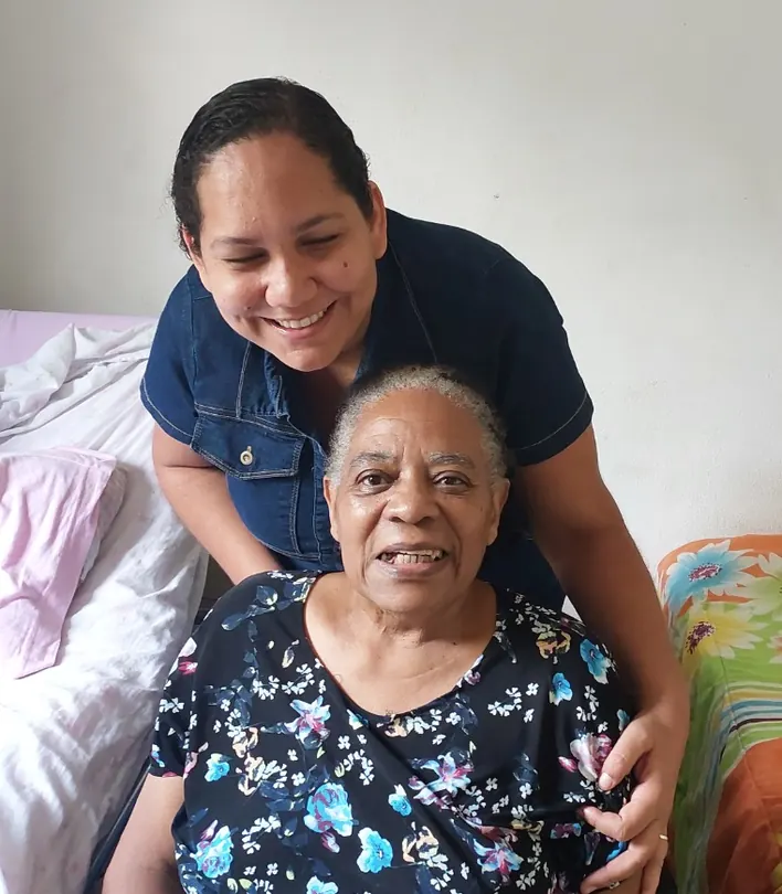 A Dida, de cabelo grisalho e vestido florido, sorri sentada enquanto uma mulher de blusa jeans a abraça por trás, também sorrindo